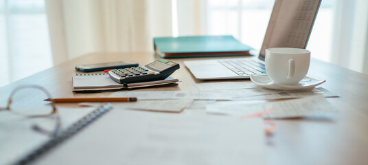 Close-up of a calculator and documents on a desk with a modern laptop in the background, next to a bright window with daylight. Business, home finances, money, and savings concept.