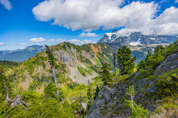 Obraz premium Rocky alpine ridge overlooking a green mountain valley with evergreen trees and distant peaks near Mount Baker in the North Cascades of Washington.