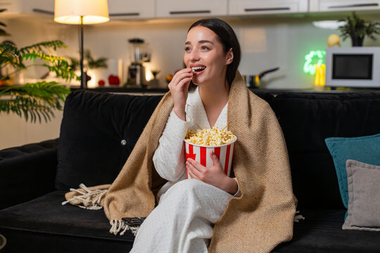 Young woman at night home watches tv or online stream laughs out loud munches popcorn happy mood late. Caucasian girl on sofa at evening comments jokes reacts to film or show friendly fun casual talk.