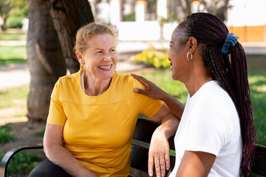 Diverse senior women friends enjoying outdoor conversation, one woman smiling and looking at her friend, feeling happy and supported by a warm touch on the shoulder