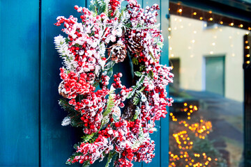 Winter charm and festive texture appear in a decorative Christmas wreath with frosted red berries, pine branches and cones hanging on a doorway, glowing softly against blurred holiday lights.