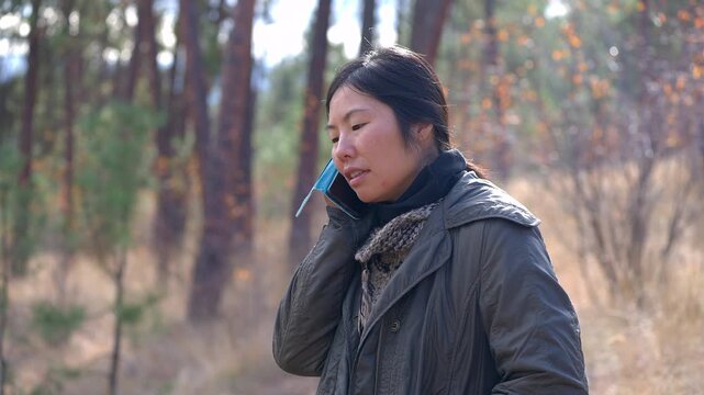 Asian woman walking in the woods in cold weather during daytime, receives and answers a phone call, speaking with caller in a friendly and familiar manner.