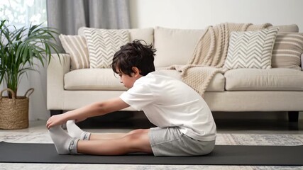 Young boy in white t-shirt and gray shorts stretches forward on a black exercise mat, in a bright living room with a beige couch and indoor plant.
