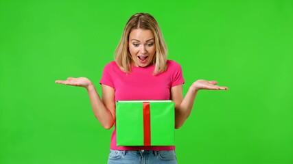 Young woman with blonde hair wearing a pink t-shirt and blue jeans, holding a green gift box with a red ribbon, looking surprised and excited against a green screen background.