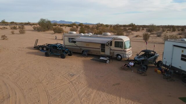 Aerial orbit of a desert camp with motorhomes, trailers, and UTVs parked in a circle.