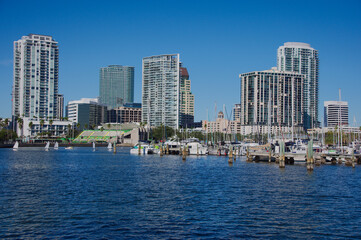 Obraz premium Sunny Waterfront Marina With High Rise Skyline And Boats In Bright Blue Sky in St. Pete, FL. Vibrant urban harbor scene featuring sailboats, a calm blue bay, and a modern skyline. A shaded concrete f