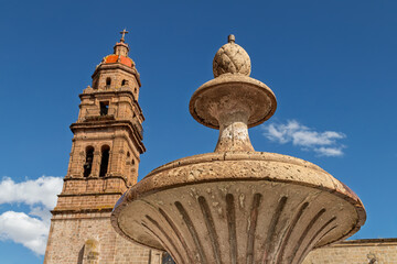 Stone fountain and colonial church bell tower under blue sky in historic center of Morelia, Mexico