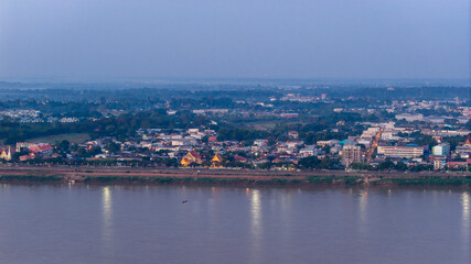Fototapeta premium Aerial drone view of Savannakhet city lights turning on at twilight. The street lights illuminate the urban landscape along the Mekong River, highlighting the city layout.