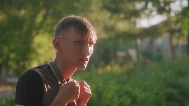 White Man Adjusting Backpack Straps And Scanning The Path, Sunlit Park Backdrop With Trees, Anxious Posture And Quick Glances Convey Hypervigilance And Restless Energy During Walk