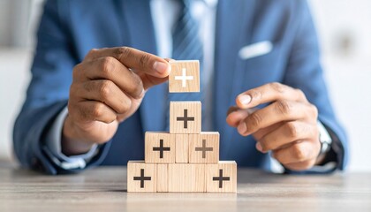Person in blue suit stacking wooden blocks with plus signs, pyramid structure, positivity and growth symbolism.