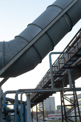 Naklejka premium Industrial duct scenery, Striking diagonal metal duct cuts through cityscape at sunset, Massive diagonal metal ventilation pipe traverses city scene with visible supports and distant structures