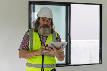 Experienced bearded construction foreman uses a digital tablet inside a new house, checking plans and managing the modern building project with confidence, precision and smart technology