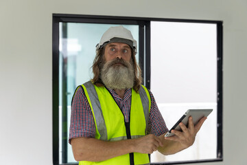 Experienced bearded construction foreman uses a digital tablet inside a new house, checking plans and managing the modern building project with confidence, precision and smart technology