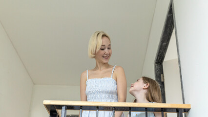Smiling mother and daughter stand on an indoor balcony of their new home, waving and pointing excitedly, sharing a warm, hopeful moment together as they imagine their future life in the house.