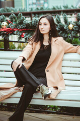 A beautiful young woman with long dark hair, wearing a black blouse, trousers, and a beige coat, sits on a park bench. Vertical photo.