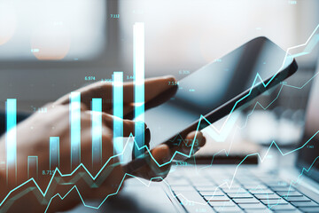 Business person analyzing digital financial charts on smartphone near laptop in office workspace with abstract glowing graph overlay effect.