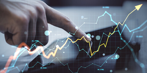 Hand interacting with tablet showing upward financial graph and currency data on transparent screen in modern digital business environment.