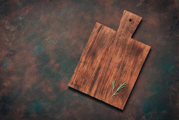 An empty wooden cutting board with a rosemary sprig on a dark grunge background. Culinary backdrop. Top view, flat lay, copy space.