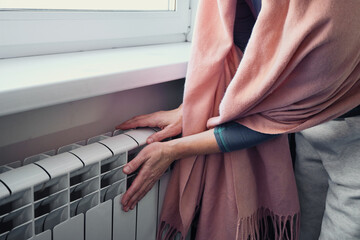 Woman warming hands on white radiator near window, feeling cold in her home