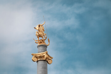 Obraz premium Tbilisi, Georgia - 3 28 2025: Golden statue of Saint George slaying the dragon atop a tall column in Liberty Square, Tbilisi, Georgia, against a partly cloudy sky. landmark and symbol of freedom.