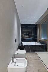 Close-up of white porcelain bidet and toilet in a modern domestic bathroom. Background shows a large bathtub. Hardwood flooring and bright lighting.