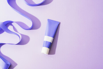 Lavender cosmetic tube on pastel purple background with flowing ribbon. Top view, copy space