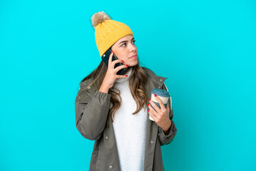 Young Italian woman wearing winter jacket and hat isolated on blue background holding coffee to take away and a mobile