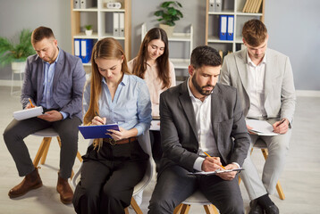 Confident coworkers being on business conference or seminar sitting on chairs indoor and writing down notes in clipboard. Group of staff working. People, engagement and work concept