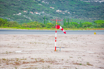 Red and white windsock indicates wind direction on a bright day at an airfield with a green hillside backdrop. Ground is sandy in the foreground next to the paved area