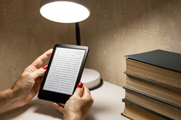  A woman reading an e-book at home. reading an e-book by lamplight. reading a book at the table. woman holding e-book. hand holding ereader. e-ink book. 