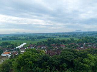 Obraz premium Peaceful aerial view of a traditional rural village and rice fields surrounded by lush greenery and distant lawu mountain under a cloudy sky. Residential area in the tropics.