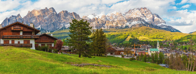 Alpine wooden chalets in Cortina d'Ampezzo town with green meadows and snow covered Dolomiti Alps mountains. Italian Dolomites in Belluno region, Italy. Alpine landscape. Travel destination