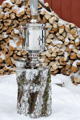 Traditional Russian samovar on a log against the background of a pile of firewood. Boiling water for tea in a classic steel samovar. Rural lifestyle in a village in Russia. Winter in the countryside.