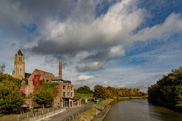 River flowing past an old brick factory chimney and church under a dramatic sky with puffy clouds.