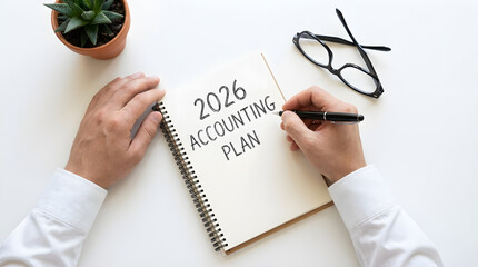 hand writing "2026 ACCOUNTING PLAN" in a notebook with a pen, black-framed glasses, and a potted plant nearby on a white table.