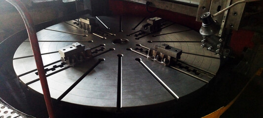 The massive, slotted rotating table of a heavy-duty vertical turning lathe, featuring several large metal chuck jaws, sits under dim industrial lighting