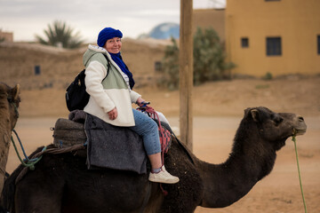 A traveler rides a camel near a desert village, dressed warmly with a blue headscarf, capturing a quiet moment of human presence within the everyday rhythm of desert life.
