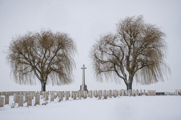 Moody winter view of a british WWI military cemetery in Belgium with cross of sacrifice and headstones covered in snow. Plugstreet battlefield site.