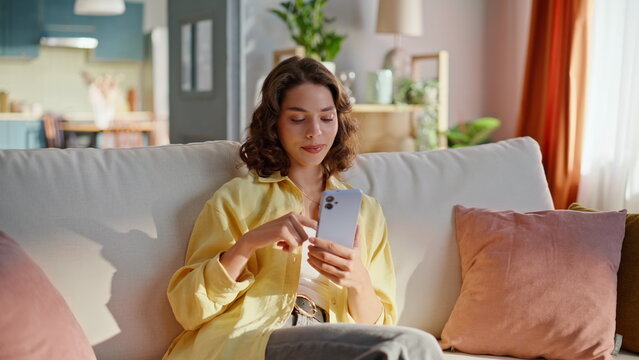 Relaxed girl scrolling cellphone at couch. Smiling woman watching news feed