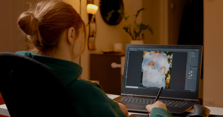 Woman is engaged in the creative process of editing images on her laptop using a graphics tablet. Her home studio is warm and inviting, showcasing her design skills.
