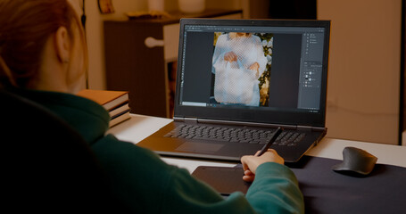 Professional photo editor working diligently on laptop, using graphics tablet for precise image retouching in cozy home studio workspace during evening hours