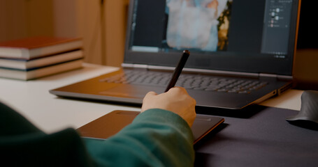 Professional graphic designer working with graphics tablet while retouching digital images on laptop in cozy home studio environment, showcasing creative workflow and digital art expertise