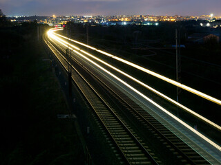 Fototapeta premium A train is traveling down a track at night. The train is very long and has a bright light on the front. The train is surrounded by a dark sky and the city lights in the background