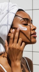 Beautiful African American woman applying face cream with closed eyes in a tiled bathroom, illuminated by artistic sunlight shadows and wearing a hair towel