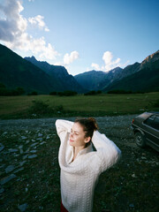 Fototapeta premium Woman wearing a chunky sweater in a mountain landscape, relaxing in a wide outdoor scene with green valley and blue sky, near a parked car during a peaceful countryside moment