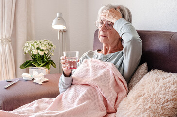 Elderly Caucasian woman with severe cold and fever symptoms, similar to seasonal flu or covid - woman on sofa at home touching forehead while taking a pill
