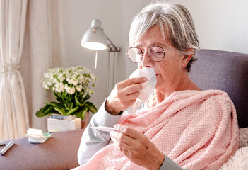 Elderly Caucasian woman with severe cold and fever symptoms, similar to seasonal flu or covid - senior woman on the couch at home checking fever on thermometer