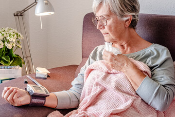Elderly Caucasian woman with severe cold and fever symptoms, similar to seasonal flu or covid - woman on the couch at home covers herself with a blanket while checking her blood pressure