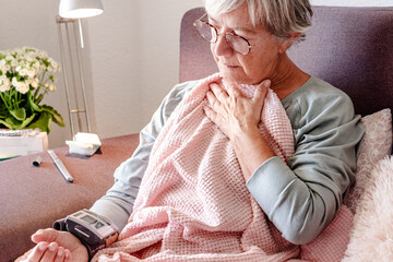 Elderly Caucasian woman with severe cold and fever symptoms, similar to seasonal flu or covid - woman on the couch at home covers herself with a blanket while checking her blood pressure
