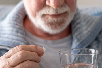 An elderly man with flu or COVID symptoms takes a pill with a glass of water. A stressed, bearded older man takes antidepressants or sedatives. Medications at work.